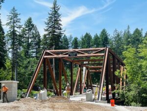 A steel truss bridge under construction, surrounded by trees, stands as a symbol of New Beginnings, with workers and orange cones lining the incomplete roadway beneath a blue sky with wispy clouds.