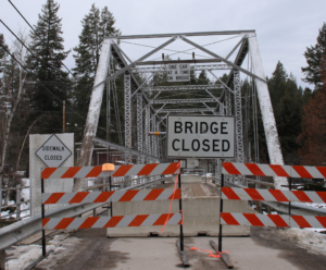A metal truss bridge blocked by striped barricades and BRIDGE CLOSED signs sits amid snow and trees, radiating cozy season vibes. A road sign above allows only one car at a time, reflecting the community spirit of shared winter paths.