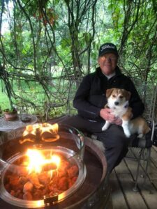 An older man wearing a cap and glasses sits on a porch during August Reflections, holding a small dog on his lap by the fire pit. Lush green vines surround them, creating a cozy, outdoor atmosphere perfect for this Season of Action.