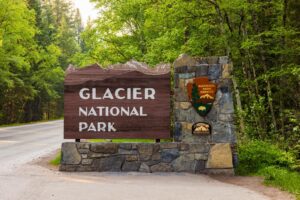 A large wooden sign reading Glacier National Park stands by a stone pillar with National Park Service emblems, surrounded by lush green trees along a paved road, welcoming visitors to new opportunities this Spring.