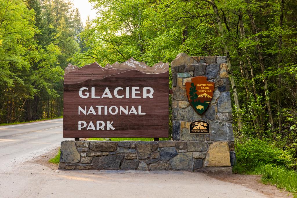 A large wooden sign reading Glacier National Park stands by a stone pillar with National Park Service emblems, surrounded by lush green trees along a paved road, welcoming visitors to new opportunities this Spring.
