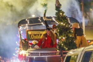 People in festive winter clothing ride a decorated holiday float with lights, a Christmas tree, and cheerful Winter Glow. One person waves a smoke flare as they pass a sign that reads BIGFORK Outdoor. The scene radiates December Real Estate magic at night.