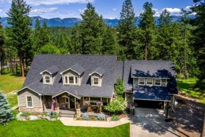 A large two-story house with gray siding, dark roof, and dormer windows sits among tall pines, its manicured lawn and front porch reflecting August Reflections, all nestled in forested mountains beneath a blue sky.