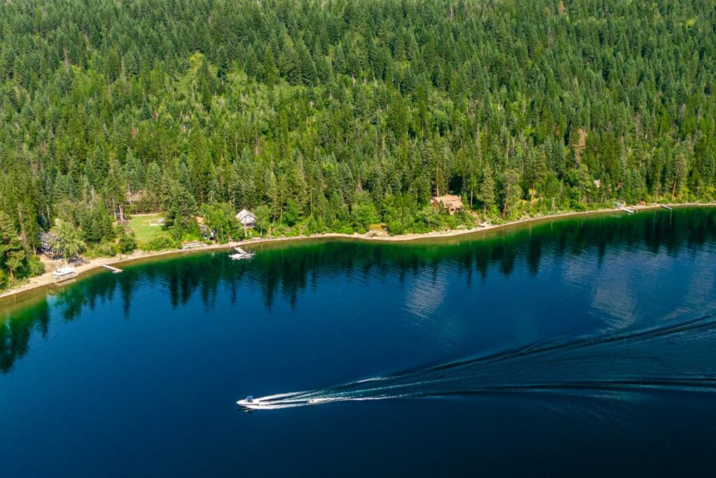 A speedboat creates white trails on a calm, blue lake bordered by dense green forest and cabins, as August keeps rolling under clear skies.