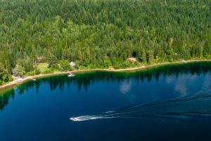 A speedboat creates a V-shaped wake on a calm, blue lake bordered by dense forest, as the changing season brings cool crisp days and hints of fall color to the trees, with a few houses and docks along the shoreline.