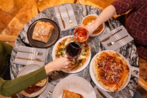 Two people clink glasses of red and rosé wine over a marble table set with pizza, a sandwich, and pasta—sharing gratitude and delicious food during the holiday season. Neatly arranged napkins and cutlery complete the festive scene.