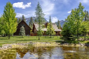 A rustic log cabin and barn surrounded by green trees sit beside a small pond, with mountain peaks and a blue sky with scattered clouds in the background.