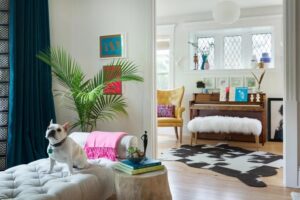 A French bulldog sits on a tufted ottoman in a colorful living room with vibrant decor, houseplants, and art—perfect for June celebrations and new beginnings. In the background, a yellow armchair and piano with a fur bench complement the bold patterned rug.