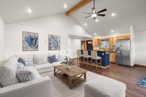 A modern open-concept living room and kitchen with a white sectional sofa, wooden coffee table, blue-accented bar, and stainless steel appliances exudes warmth—perfect for gathering in gratitude during the holiday season. Botanical art and wood finishes complete the look.