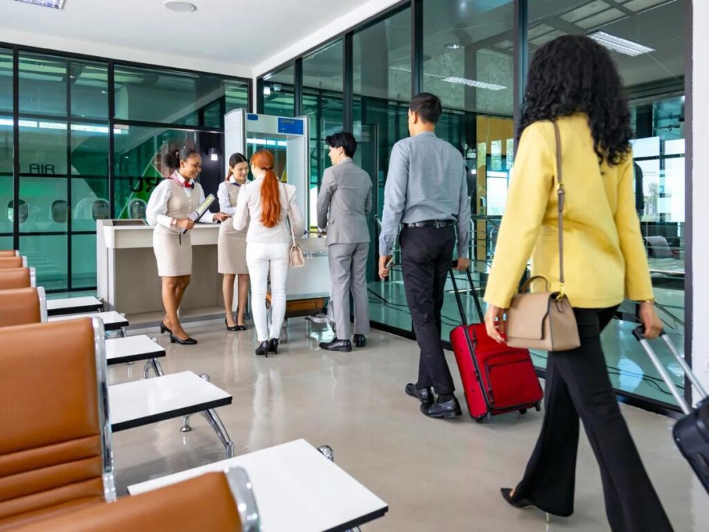 Five travelers with luggage stand in line at an airport boarding gate, interacting with two staff members. As Spring brings new opportunities, empty chairs await in the lounge, and large windows reveal part of an airplane outside.