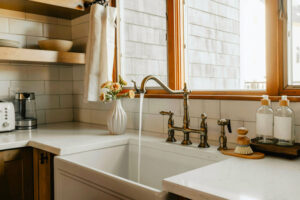 A farmhouse-style kitchen sink with water running from a brass faucet, surrounded by soap dispensers, a scrub brush, and a vase with flowers on a white countertop—capturing that Winter Glow beneath a wooden window.