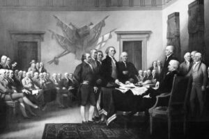 A group of men in 18th-century clothing gather around a table in a formal room, some standing to sign a document while others observe. Listings of patriotic flags hang on the wall behind them, marking an important moment in history.
