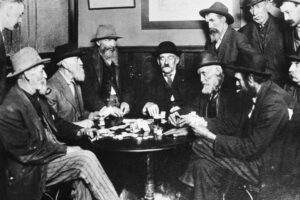 A group of older men in hats and suits sit and stand around a small round table, playing cards and smoking with intention, in a dimly lit room with wood-paneled walls.