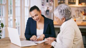 A young woman in business attire sits at a table with an older woman, smiling and discussing documents—marking new beginnings. A laptop is open on the table in a bright, cozy home setting.