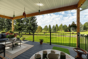 A covered patio with outdoor seating, potted plants, and a table overlooks a lush, green lawn with trees under a partly cloudy spring sky. String lights hang from the wooden beams above the railing, adding a touch of charm to the season.