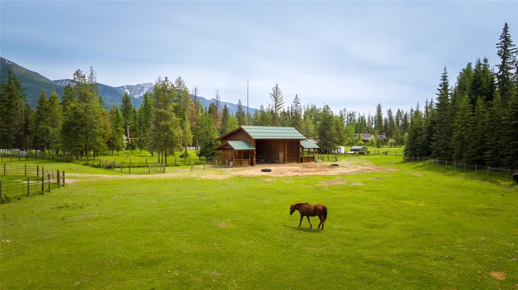 A brown horse grazes on green grass in front of a wooden barn, surrounded by fences, tall pine trees, and distant mountains under a cloudy sky, creating a peaceful scene perfect for a Merry Christmas or Bright New Year greeting.