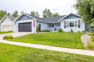 A single-story blue house with white trim, a dark garage door, and an American flag stands proudly in Flathead Valley, its well-maintained lawn and landscaping inviting you to enjoy the bright day of the Spring Market season.