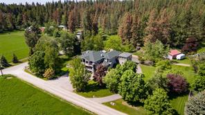 Aerial view of a large house surrounded by trees and greenery, with a paved road in front and forested area in the background—perfect for the HOT Summer Listing Season. Other small buildings are visible nearby.