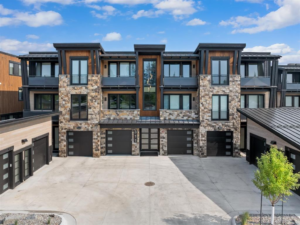 Modern three-story townhouse with stone and wood exterior, large windows, balconies, and multiple garages on the ground level. The stylish design reflects intention and new beginnings, with a driveway and small tree under a blue sky with clouds.