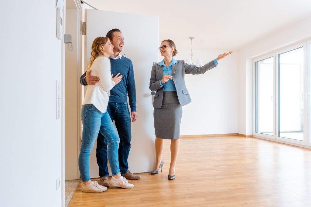 A smiling real estate agent from Life Listings shows an empty, bright room with wooden floors and large windows to a happy couple standing at the door.