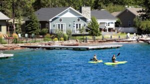 Two people kayak on a blue lake in Flathead Valley, passing a gray house with a stone chimney and white trim, surrounded by trees and docks—an inviting scene as the Spring Market arrives.