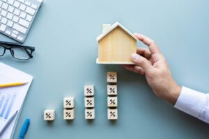 A hand holds a small wooden house above stacked percentage blocks, forming a staircase—a symbol of new beginnings and intention. Nearby rest a keyboard, glasses, charts, pen, and pencil on a light blue desk.