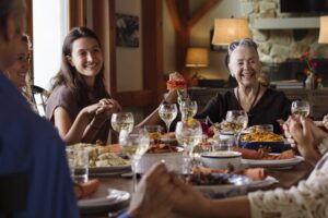 A group of people of different ages sit around a dining table, sharing a meal and smiling. The table is set with food, wine glasses, and plates. The scene feels warm and cheerful, filled with gratitude during the holiday season in a cozy home.
