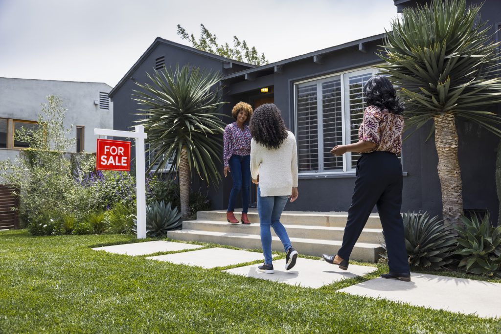 Three women approach the entrance of a modern house with a For Sale sign in the yard on a sunny day, capturing the spirit of August Reflections and hinting at new beginnings during this Season of Action.
