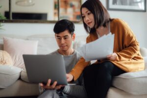 A man and woman sit together in a living room, looking at a laptop. The woman, holding papers, points at the screen as they review documents—perhaps planning for Fall in Bigfork or preparing for September in Bigfork.