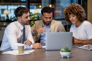 Three business professionals, two men in suits and a woman in a white top, gather around a laptop, discussing July Journeys. Papers, a tablet, and a coffee cup are spread across the table as they review important details together.