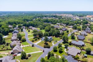 Aerial view of a suburban neighborhood with tree-lined streets, green lawns, and houses spaced apart, surrounded by dense woods under the steady momentum of February light and a clear sky.