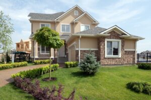 Two-story suburban house with beige walls, stone accents, and a gray roof, surrounded by a well-manicured lawn and blooming homes of trees and shrubs under a partly cloudy sky.