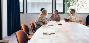 Four people sit around a long conference table in a modern office, engaged in discussion. As laptops and notebooks rest on the table, it's clear the market is heating up—large windows and a potted plant complete the scene.