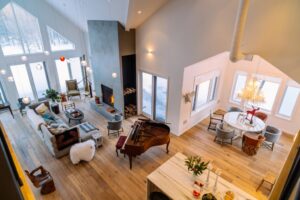 Spacious, modern living and dining area with high ceilings, large windows, a cozy fireplace, a grand piano, contemporary furnishings, and wooden floors—perfect for enjoying winter real estate and snowy views through the windows.