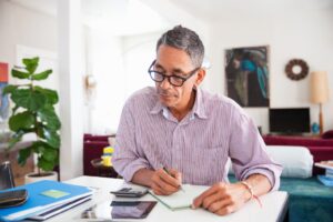 A middle-aged man with glasses sits at a table in a bright living room, writing in a notebook. Surrounded by papers, a calculator, and folders, he appears focused—ready to seize new opportunities this Spring.