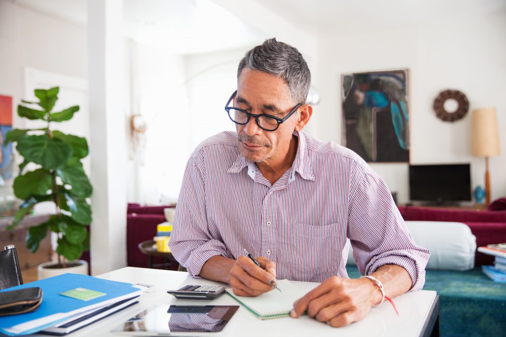 A middle-aged man with glasses sits at a table in a bright living room, writing in a notebook. Surrounded by papers, a calculator, and folders, he appears focused—ready to seize new opportunities this Spring.