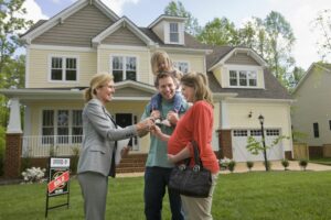 A real estate agent hands keys to a smiling family—parents with a young child on the father’s shoulders—standing in front of a large suburban house with a “sold” sign in the yard as the market is heating up.