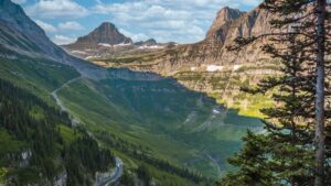 A scenic mountain landscape with a winding road, lush green valleys, rocky peaks, patches of snow, and a partly cloudy blue sky, viewed through pine trees in the foreground—perfectly capturing the vibrant beauty of fall in this October update.