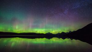 Vivid green and purple aurora borealis lights stretch across a star-filled night sky, reflecting on a calm lake with silhouetted mountains—a mesmerizing winter real estate view perfect for December.