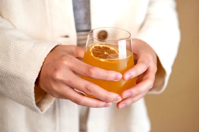 A person wearing a cream-colored jacket holds a round glass filled with an orange drink, garnished with a dried citrus slice—perfect for toasting a Bright New Year.