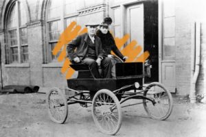 A black-and-white photo of a man and woman in an old-fashioned, open-top car with large spoked wheels, parked before a brick building; orange brush strokes evoke the warmth of February Light behind them.