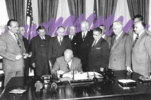 A man sits at a large desk signing a document while ten men stand around him, observing. The formal office, adorned with flags and heavy curtains, evokes the crisp transition from Summer to Fall in Bigfork.