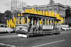 A vintage San Francisco cable car travels down a street; the photo is black and white, with yellow squiggles drawn in the background behind the cable car, evoking a sense of steady momentum through the cityscape.
