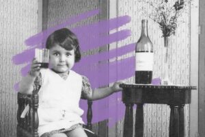 A young child sits on a wooden chair, holding up a glass and smiling. Next to the child, a bottle rests on a small round table with flowers, while wallpaper and a purple paint streak hint that love is in the air.