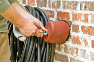 A person lifts a red foam cover to access an outdoor faucet on a brick wall, preparing for winter real estate needs, with a coiled garden hose hanging nearby.