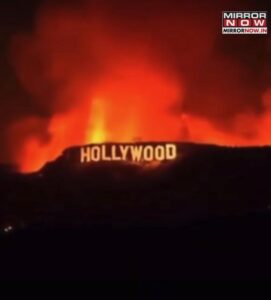 The iconic Hollywood sign is illuminated at night with large flames and thick smoke in the background, suggesting a wildfire on the hillside after a Winter Pause. The Mirror Now news logo appears in the top right corner.