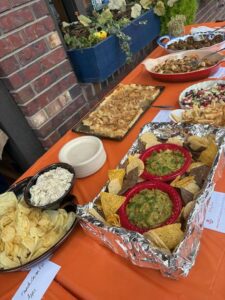 An orange tablecloth holds several dishes, including chips with guacamole, dip with potato chips, a baked pasta dish, and salad. Paper labels are by each dish as community lights twinkle against a brick wall and potted plants in the background.