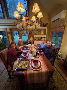 Five smiling adults sit around a warmly lit dining table set with plates, bowls, candles, and food, sharing gratitude inside a cozy room with large windows and rustic decor during the holiday season.