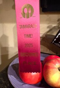 A red ribbon reading Tamaracka Time! 2025 Second Place stands upright, supported by an apple on a plate with two others, glowing softly like community lights. The ribbon features a fork and spoon logo at the top.