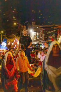People dressed in colorful costumes walk in a festive nighttime parade, bubbles drifting through the Winter Glow and bright holiday lights. A person wearing a crown stands on a float behind others, adding magic to this December Real Estate celebration.